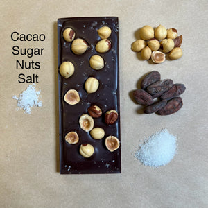 A bar of dark chocolate with hazelnuts on top, accompanied by a small pile of salt crystals and whole hazelnuts to the side.
